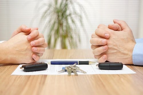 Close up of a pair of hands across from each other on a table, resting on a piece of paper that has a pen, two key fobs, and keys.